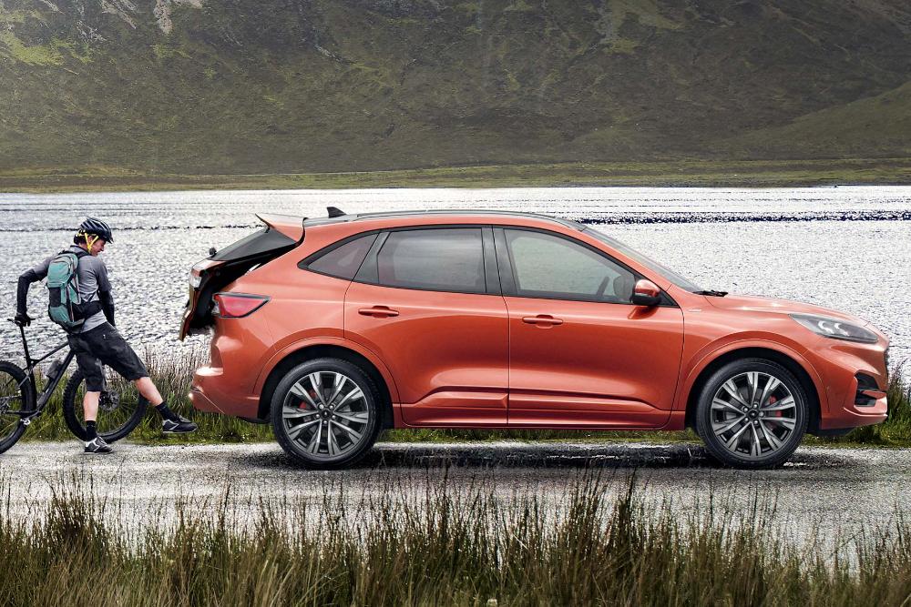 Een oranje Ford Kuga van opzij, geparkeerd bij een meer, met een fietser die spullen in de open kofferbak legt.