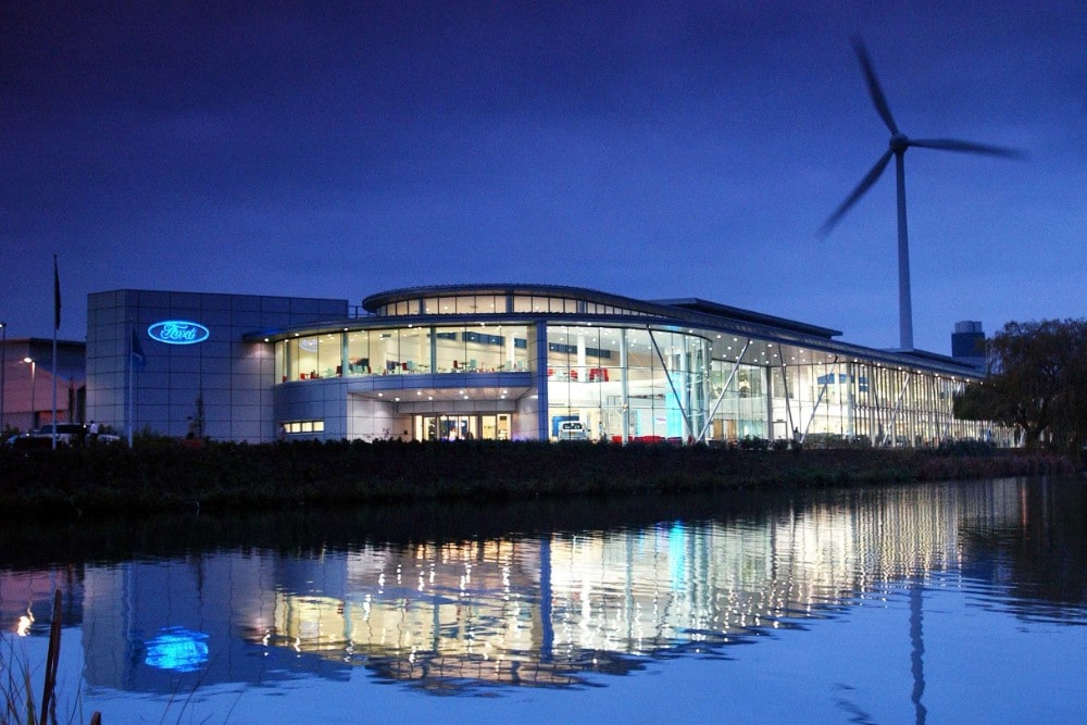 Een verlicht Ford-gebouw bij schemering, met een windturbine die weerspiegelt in het water op de voorgrond.