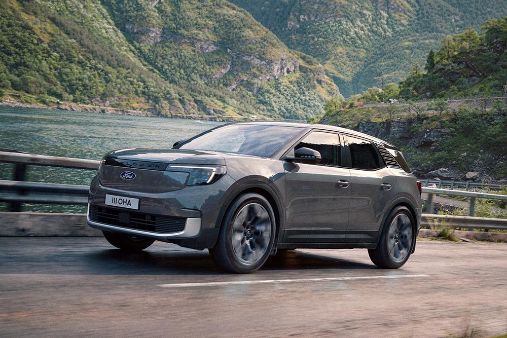 Grijze Ford Explorer rijdend op een bergweg langs een fjord met beboste bergen.