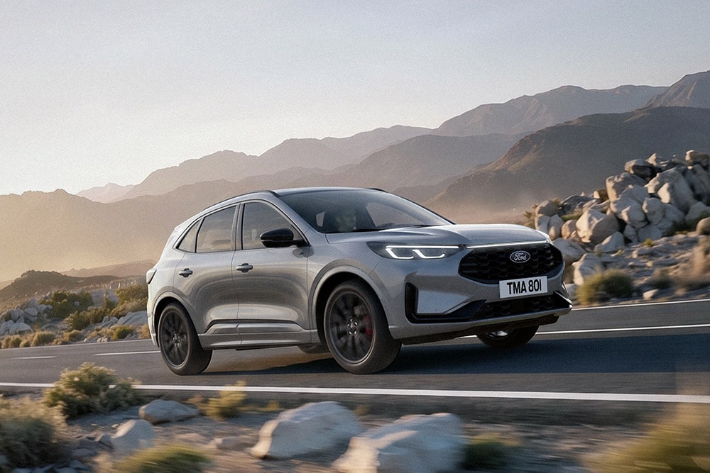 Zilvergrijze Ford Kuga rijdend op een bergweg tussen rotsen en bergketens.