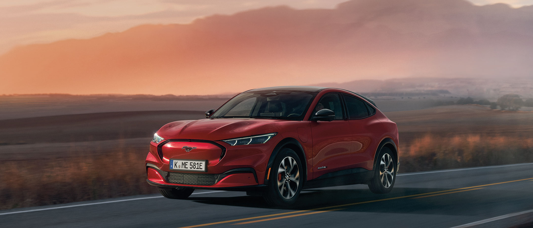 A red Mustang Mach-E on a road, with the sunset behind it.