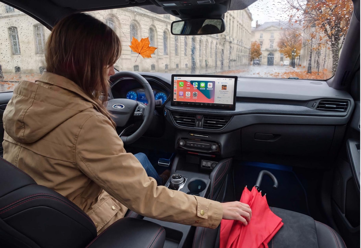 Een binnenopname van een Ford-auto in de herfst. Een vrouw in een bruine jas zit op de bestuurdersstoel en reikt naar een rode paraplu op de passagiersstoel.