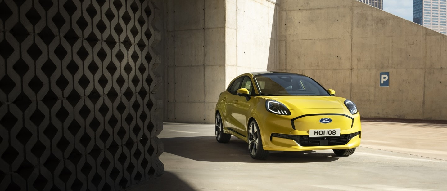 Een felgele Ford Puma Gen-E staat geparkeerd op een betonnen oppervlak onder een moderne, hoekige betonnen architectuur.