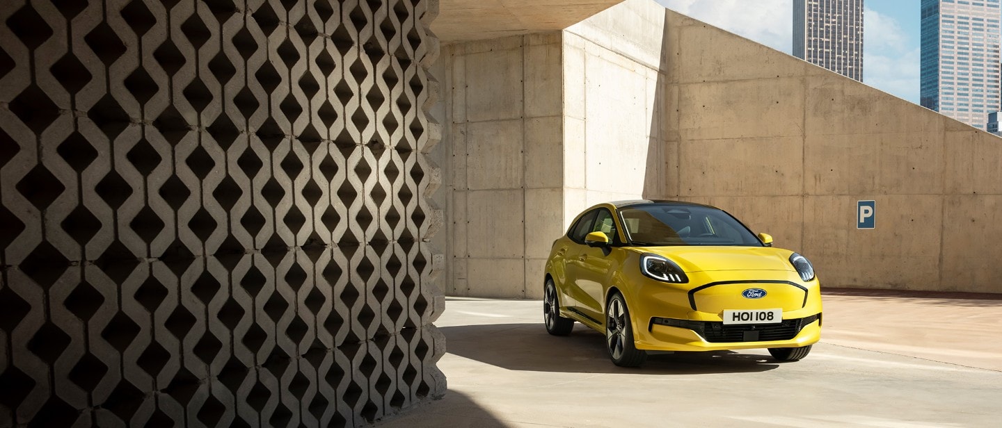 Een felgele Ford Puma Gen-E staat geparkeerd op een betonnen oppervlak onder een moderne, hoekige betonnen architectuur.