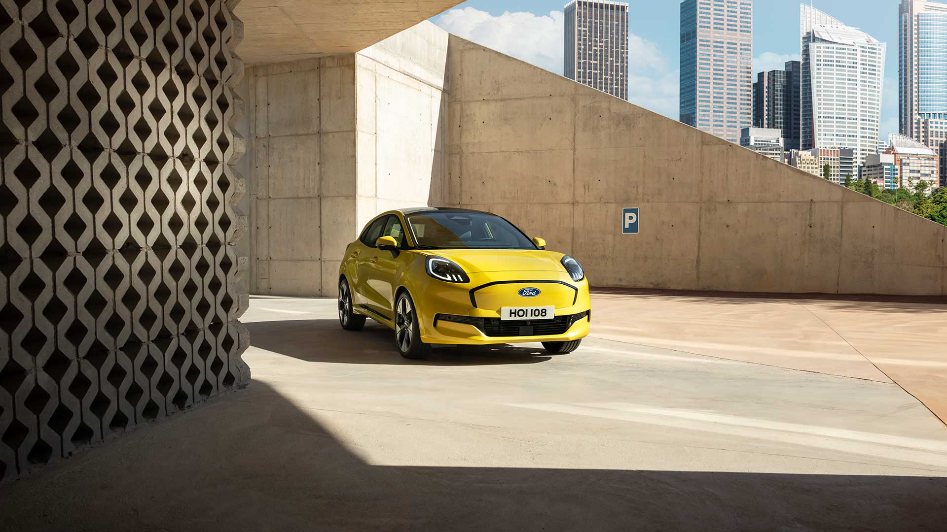 Een felgele Ford Puma Gen-E staat geparkeerd op een betonnen oppervlak onder een moderne, hoekige betonnen architectuur.