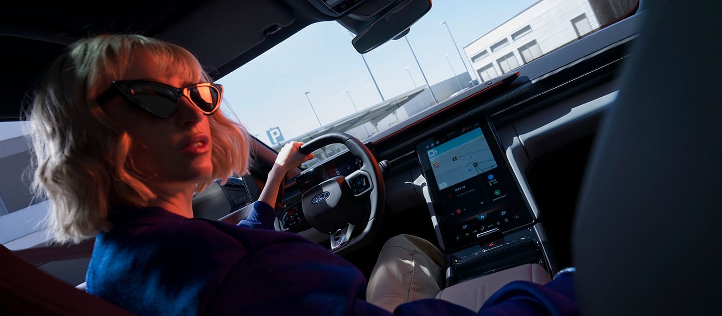Een vrouw zit op de bestuurdersstoel van de elektrische Ford Capri, en toont ook het grote 14,6" SYNC Move touchscreen.