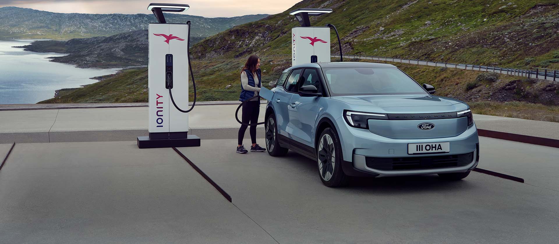 Een vrouw sluit een blauwe, 100% elektrische Ford Explorer aan op een openbaar laadstation met een kabel. De auto staat geparkeerd bij bergen en water. 