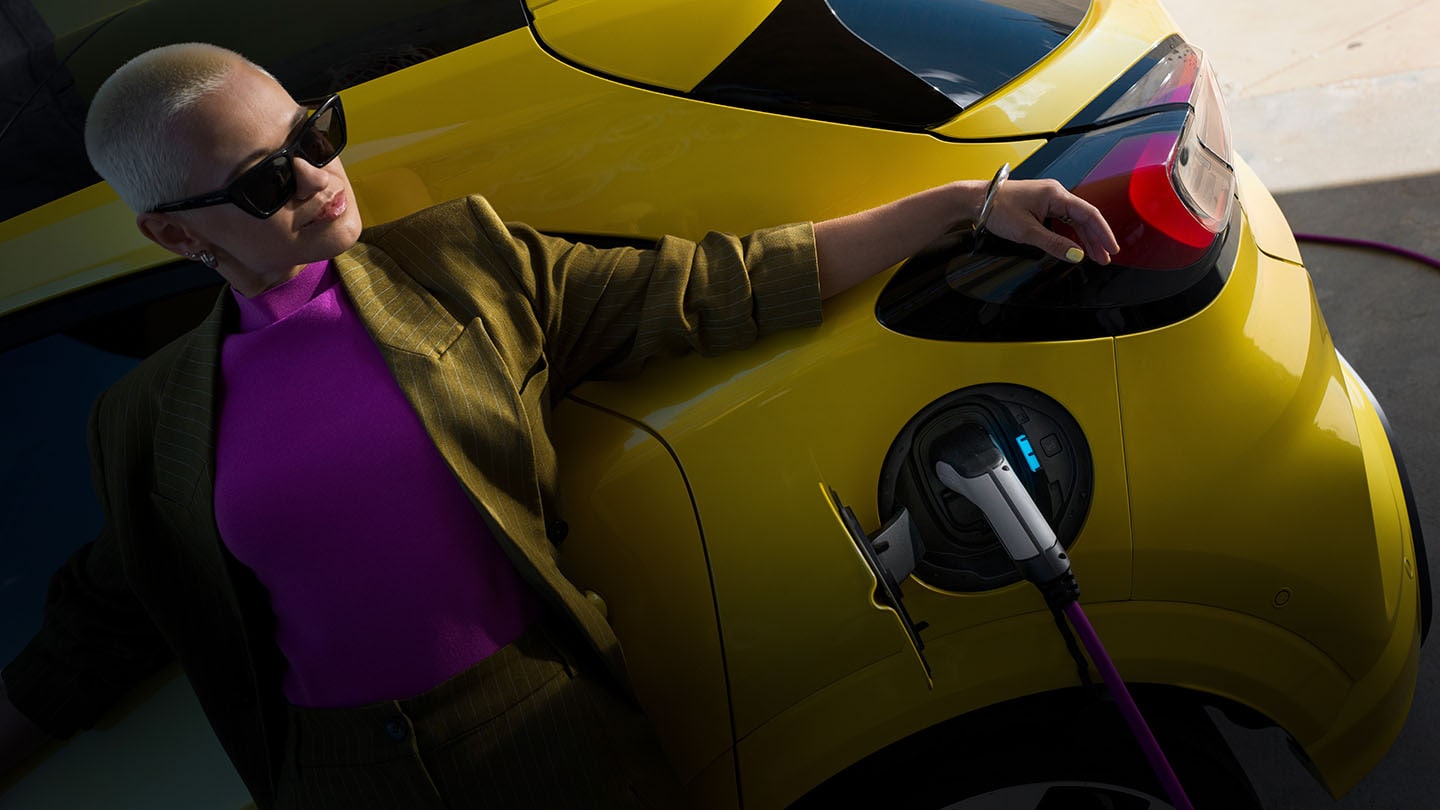 Een vrouw leunend tegen een gele elektrische Ford Capri die is aangesloten op een EV-oplader.