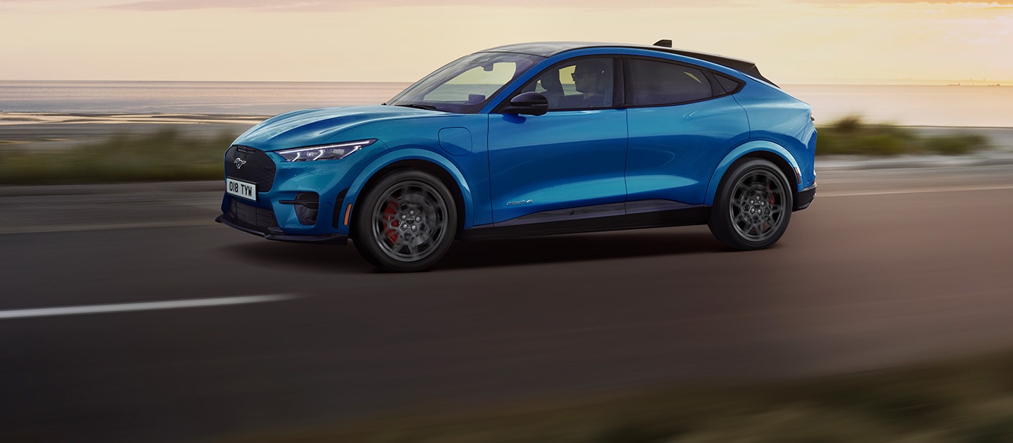 Een blauwe elektrische Ford Mustang Mach-E rijdt langs een kustweg bij zonsondergang.