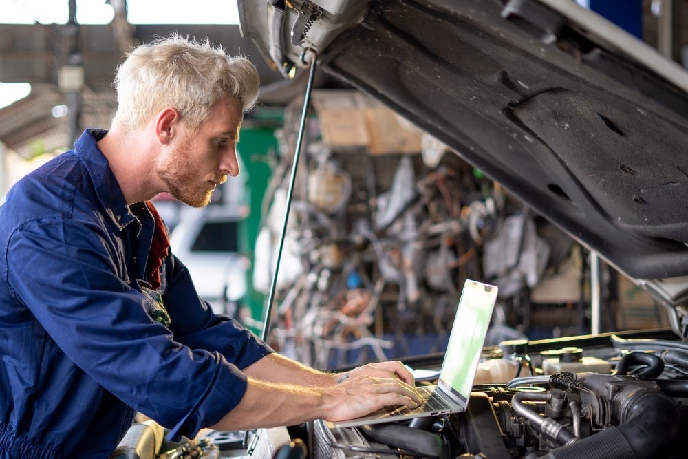 Een technicus in een blauw uniform gebruikt een laptop terwijl hij werkt aan een automotor met open motorkap.