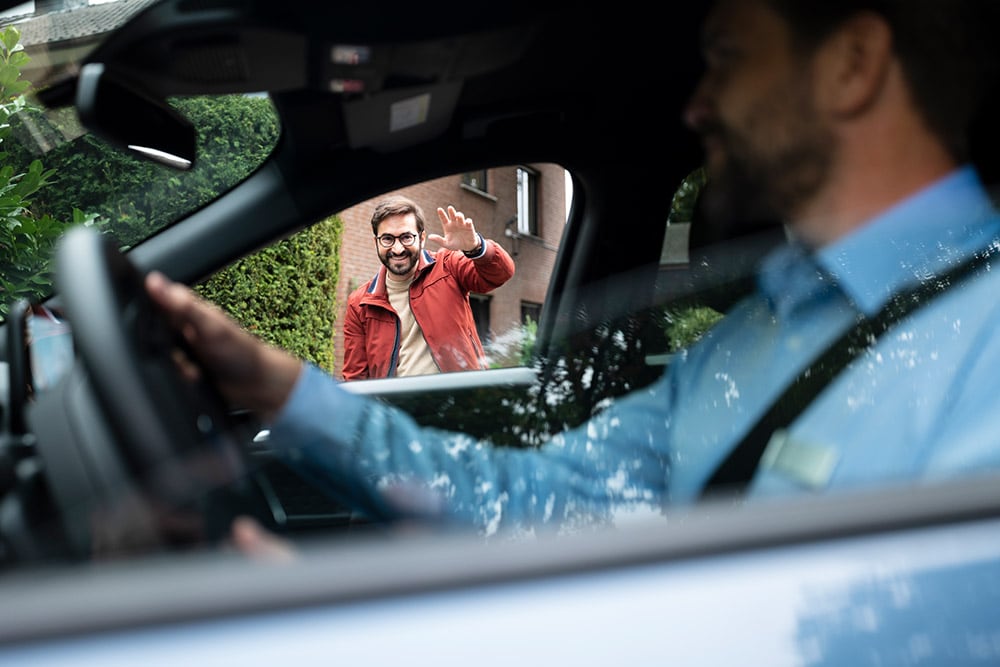 Een man aan het stuur van een auto groet een andere glimlachende man buiten.