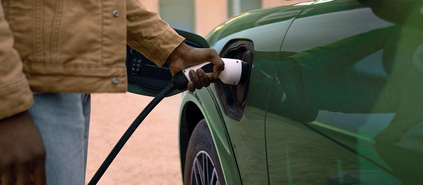 Een persoon in een crèmekleurige jas die een elektrische oplaadkabel aansluit op zijn groene elektrische Ford Mustang Mach-E.