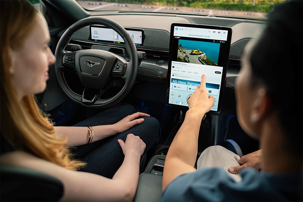 Close-up op het dashboard van een Mustang Mach-E, een chauffeur wijst naar het touchscreen.