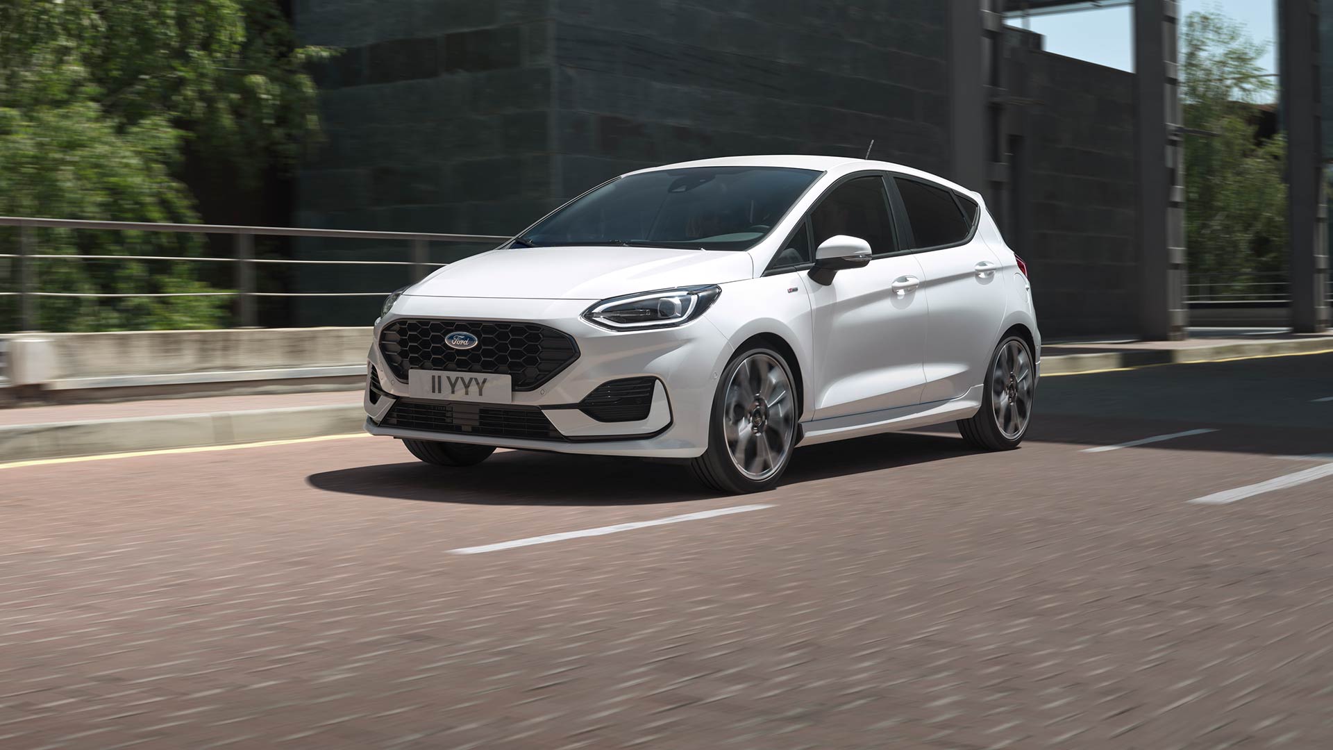 Witte Ford Fiesta rijdt op een stadsweg, groen en gebouwen op de achtergrond.