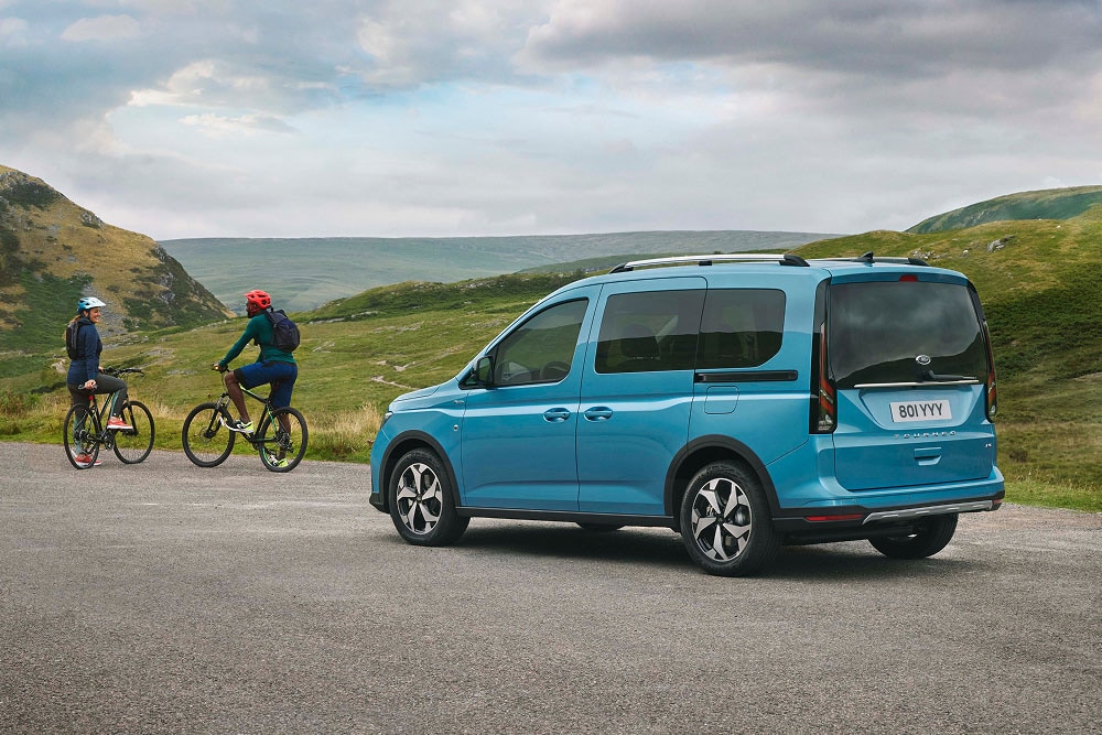 Een blauwe Ford Tourneo Connect staat geparkeerd langs een weg in een heuvelachtig landschap, met twee fietsers op de achtergrond