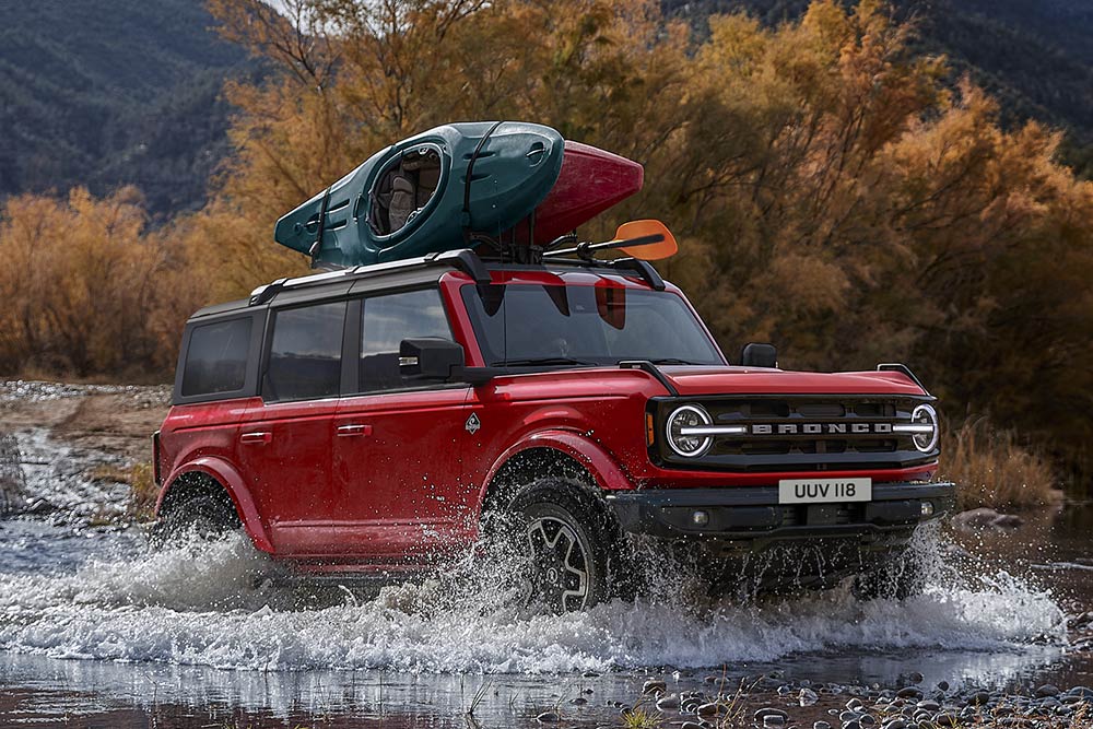 Zijaanzicht van een rode Ford Bronco door ondiep water in een bergachtige omgeving.