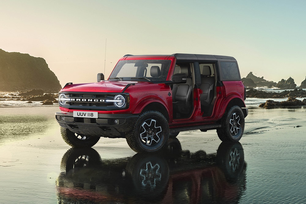 Zijaanzicht van een rode Ford Bronco, met verwijderde deuren, geparkeerd op een nat zandstrand.
