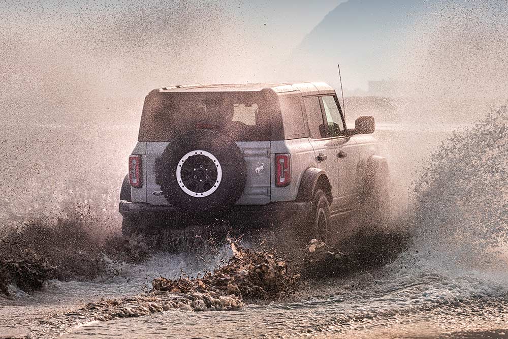 Een achteraanzicht van een zilveren Ford Bronco door water op een strand.