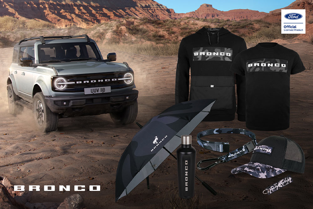 Ford Bronco-collectie met zwart T-shirt, jas, polo, pet, muts en regenrugzak, en een grijze Bronco op de achtergrond.