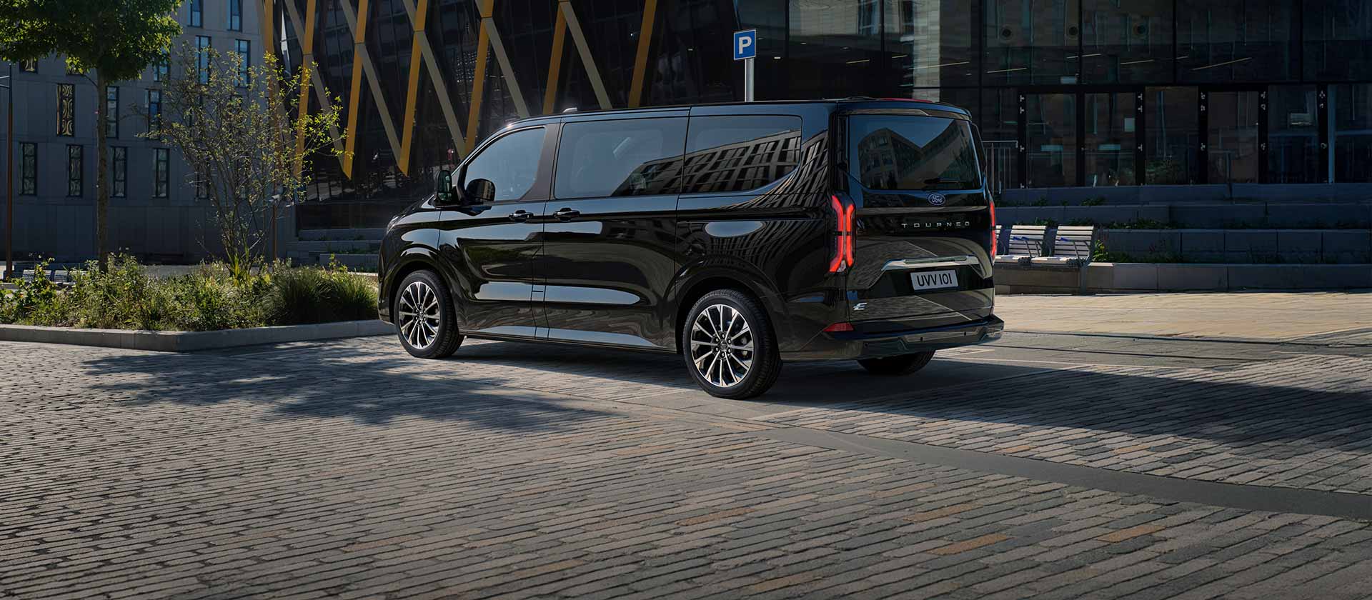 Een zwarte Ford E-Tourneo Custom staat geparkeerd met de zijkant en achterkant voor een modern gebouw.
