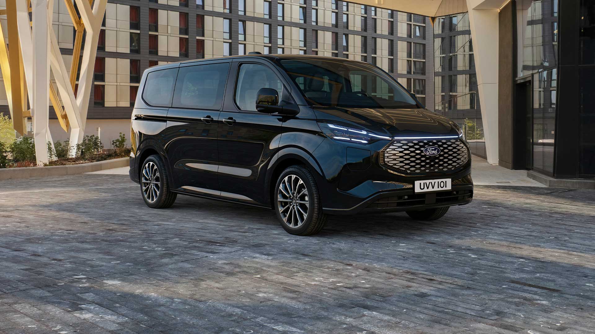 Een zwarte Ford E-Tourneo Custom staat geparkeerd voor een modern gebouw.