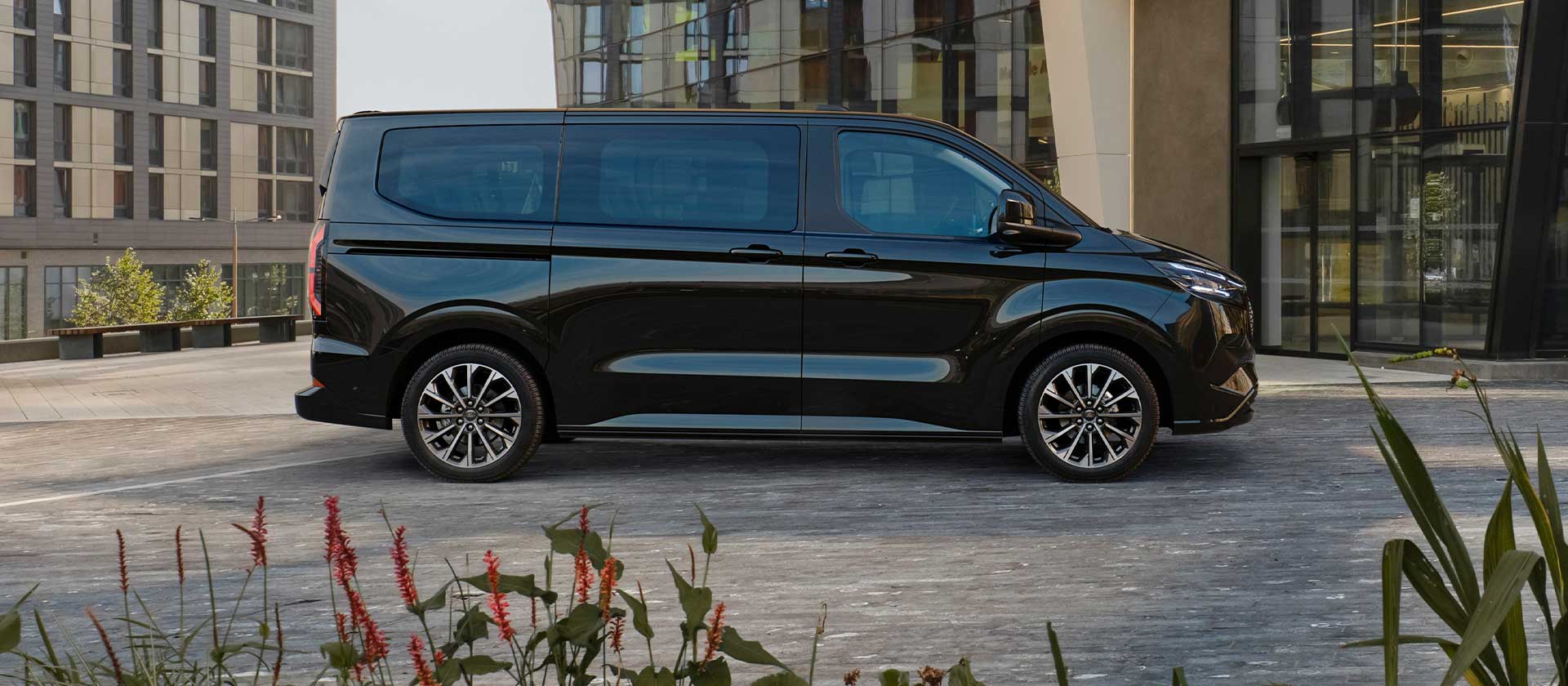 Een zwarte Ford E-Tourneo Custom staat geparkeerd voor een modern gebouw.