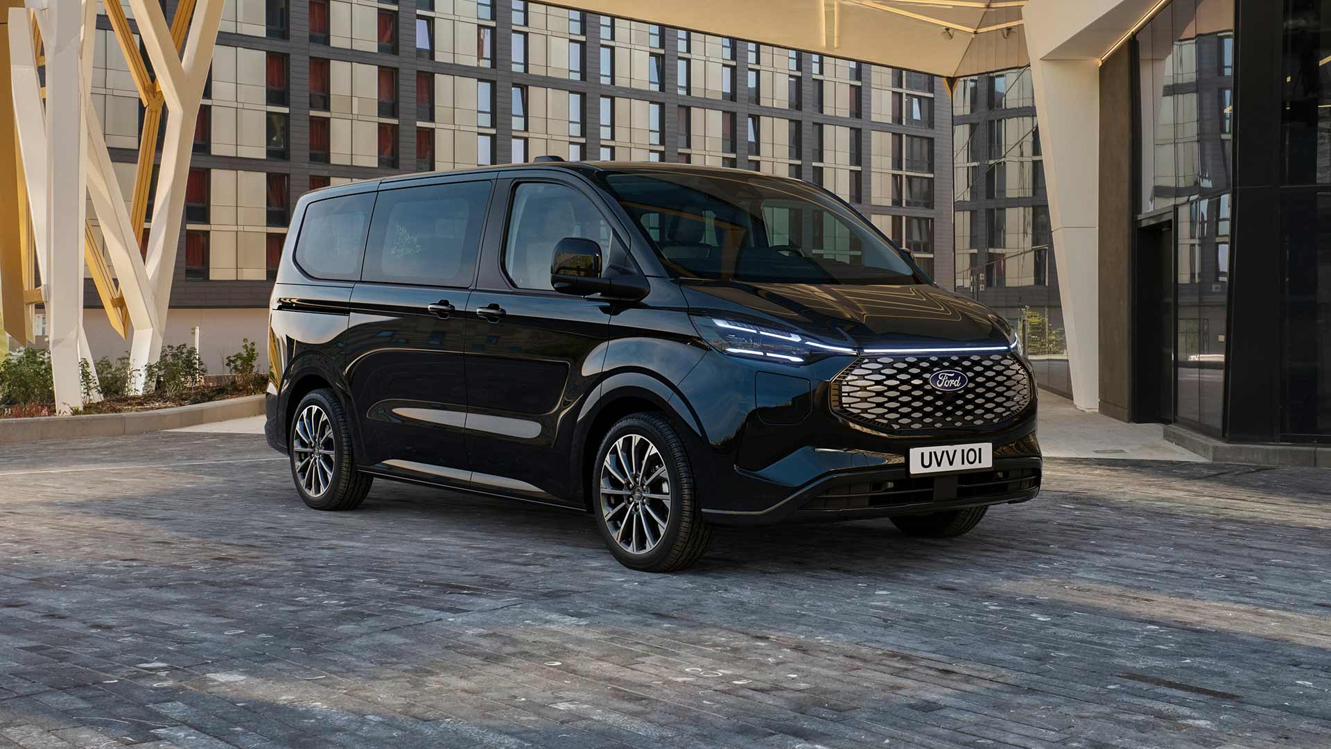 Een zwarte Ford E-Tourneo Custom staat geparkeerd voor een modern gebouw.