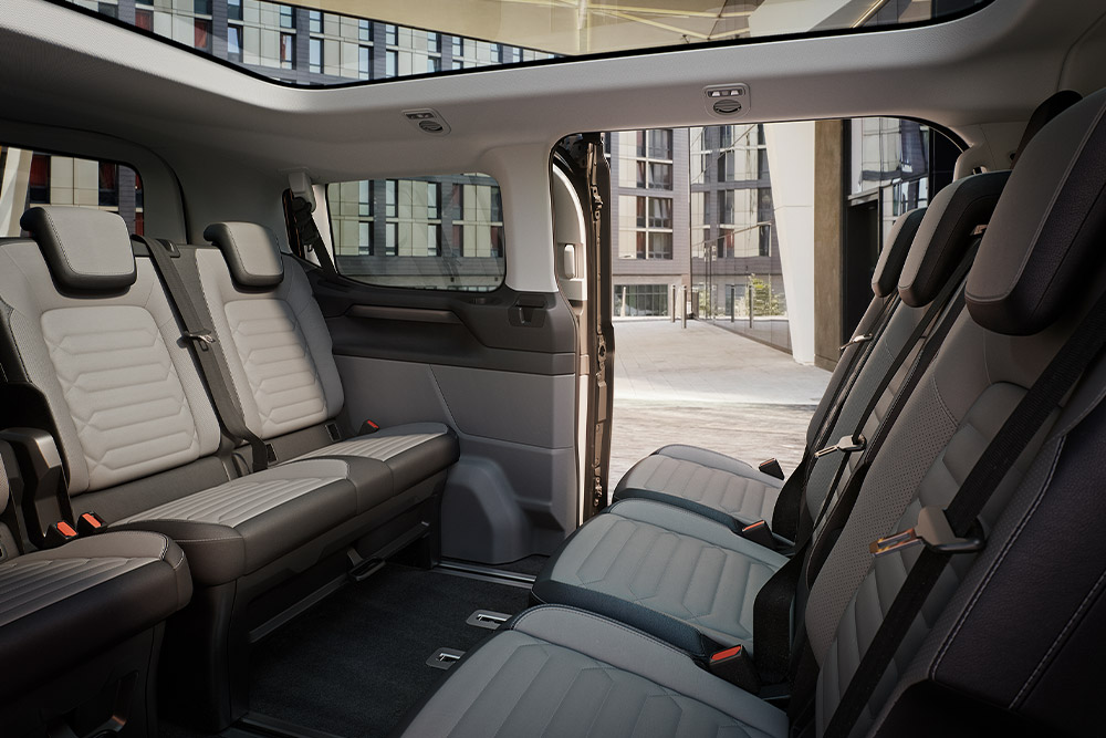 Binnenaanzicht van de Ford Tourneo Custom, met meerdere rijen grijze passagierszetels en een panoramisch zonnedak.