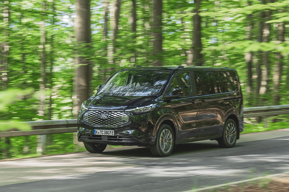 Een zwarte Ford E-Tourneo Custom rijdt langs een met bomen omzoomde weg.