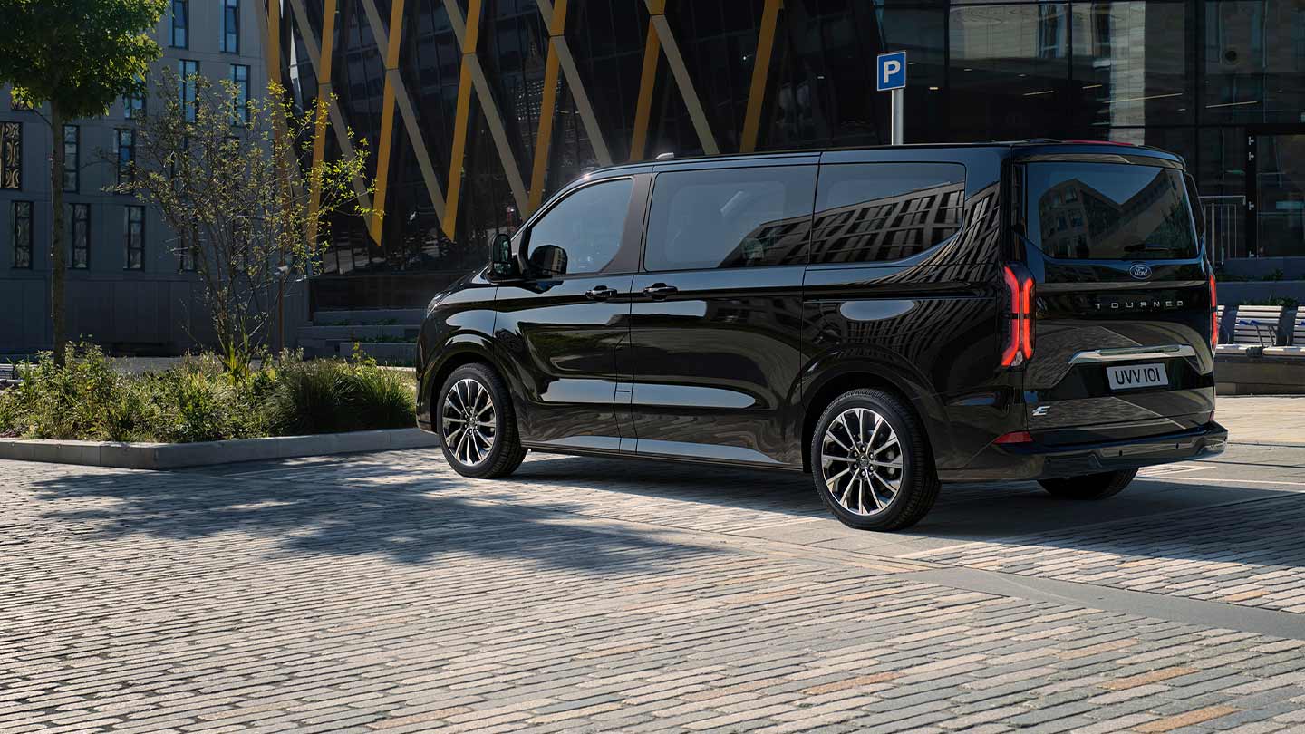 Een zwarte Ford E-Tourneo Custom staat geparkeerd met de zijkant en achterkant voor een modern gebouw.