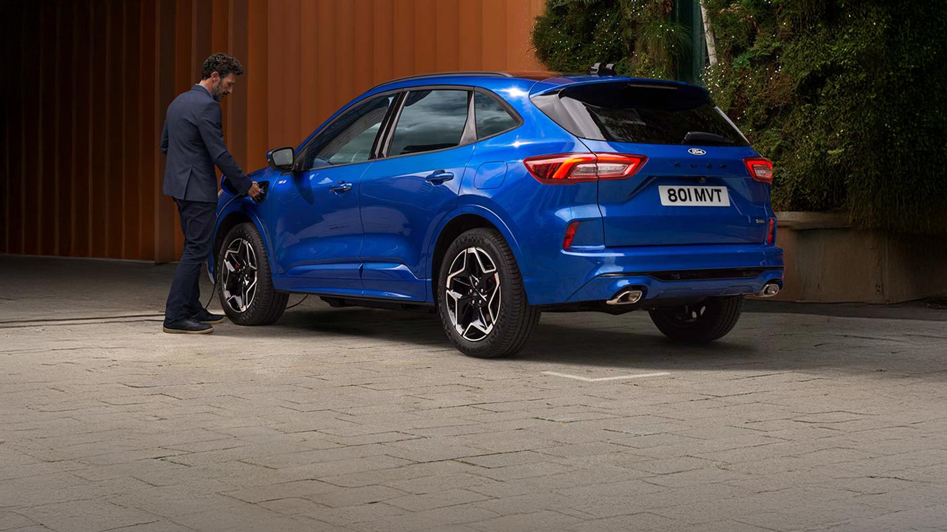 Achteraanzicht van een blauwe Ford Kuga die door iemand wordt opgeladen in een stedelijke omgeving.