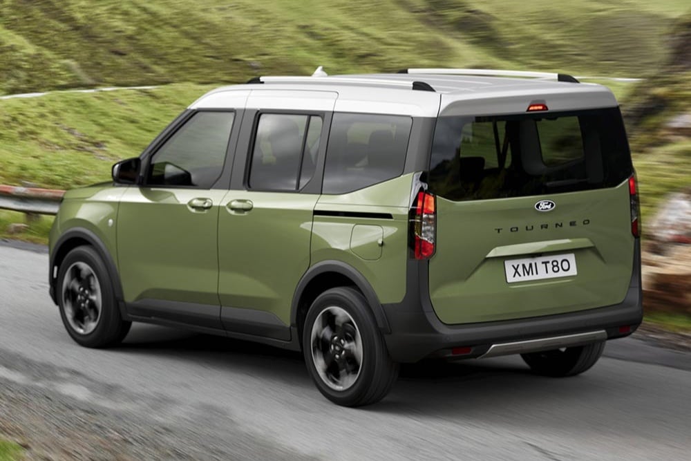 Een groene Ford Tourneo Courier van achteren in een driekwarthoek gezien, rijdend op een kronkelende weg met groene heuvels.