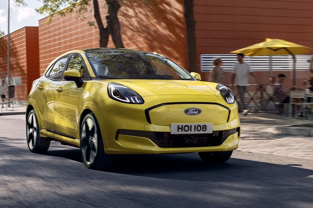 Vooraanzicht van een gele elektrische Ford Puma Gen-E® die langs een stadsweg rijdt.