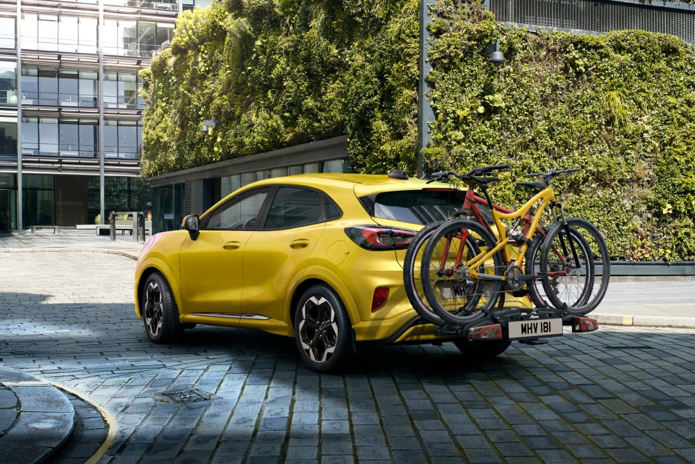 Gele Ford Puma Gen-E geparkeerd in een stedelijke omgeving met twee fietsen op een fietsendrager achterop.