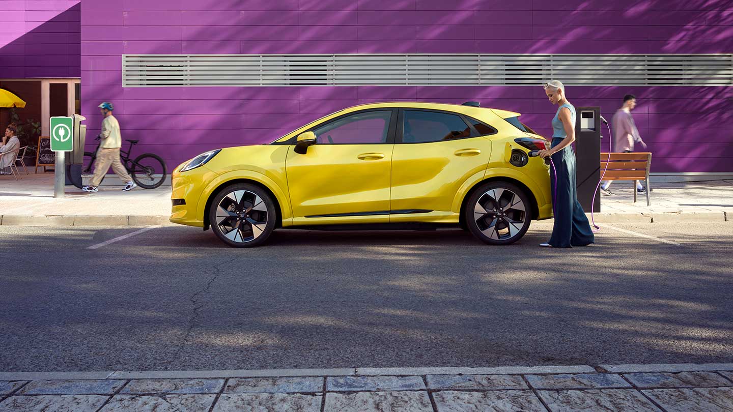 Zijaanzicht van een gele elektrische Ford Puma Gen-E® aangesloten op een openbaar laadstation terwijl hij geparkeerd staat.