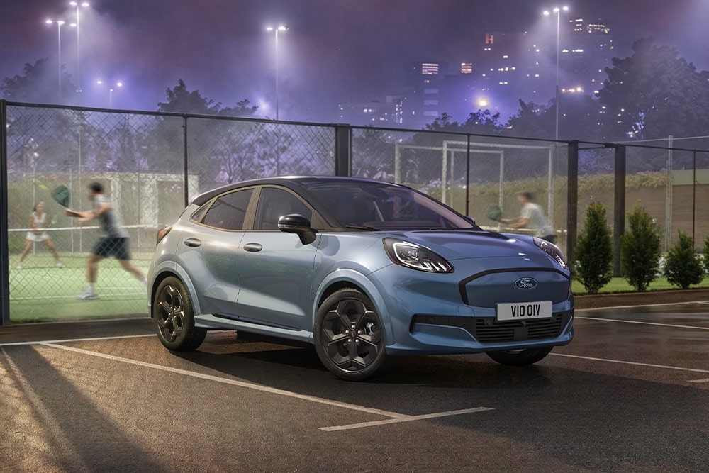 Een blauwe Ford Puma Gen-E geparkeerd naast een tennisbaan, met stadslichten op de achtergrond.