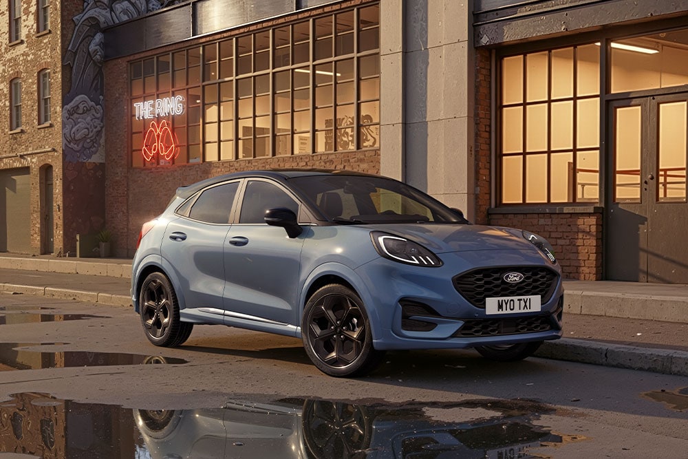 Een blauwe Ford Puma geparkeerd op een stadsstraat bij zonsondergang, met plassen die de gebouwen weerspiegelen.
