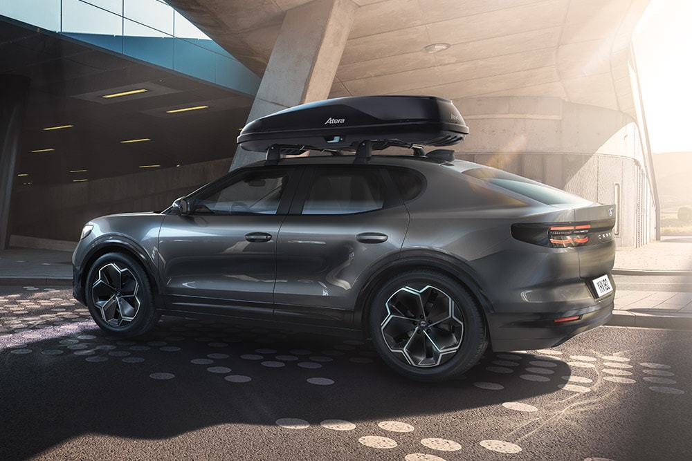 3/4 zijaanzicht van een rode Ford Capri met dakkoffer, geparkeerd onder een moderne gebouwstructuur.