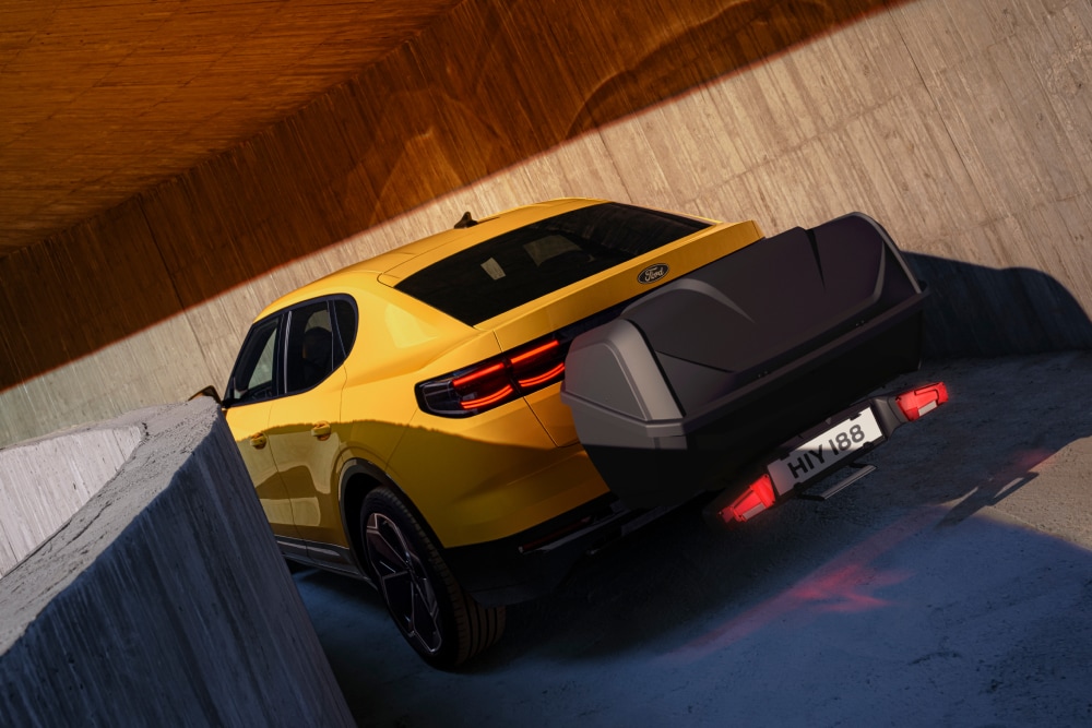 Gele Ford Capri geparkeerd in een moderne betonnen constructie, met een bagagebox achterop die aan een bagageboxdrager is bevestigd.