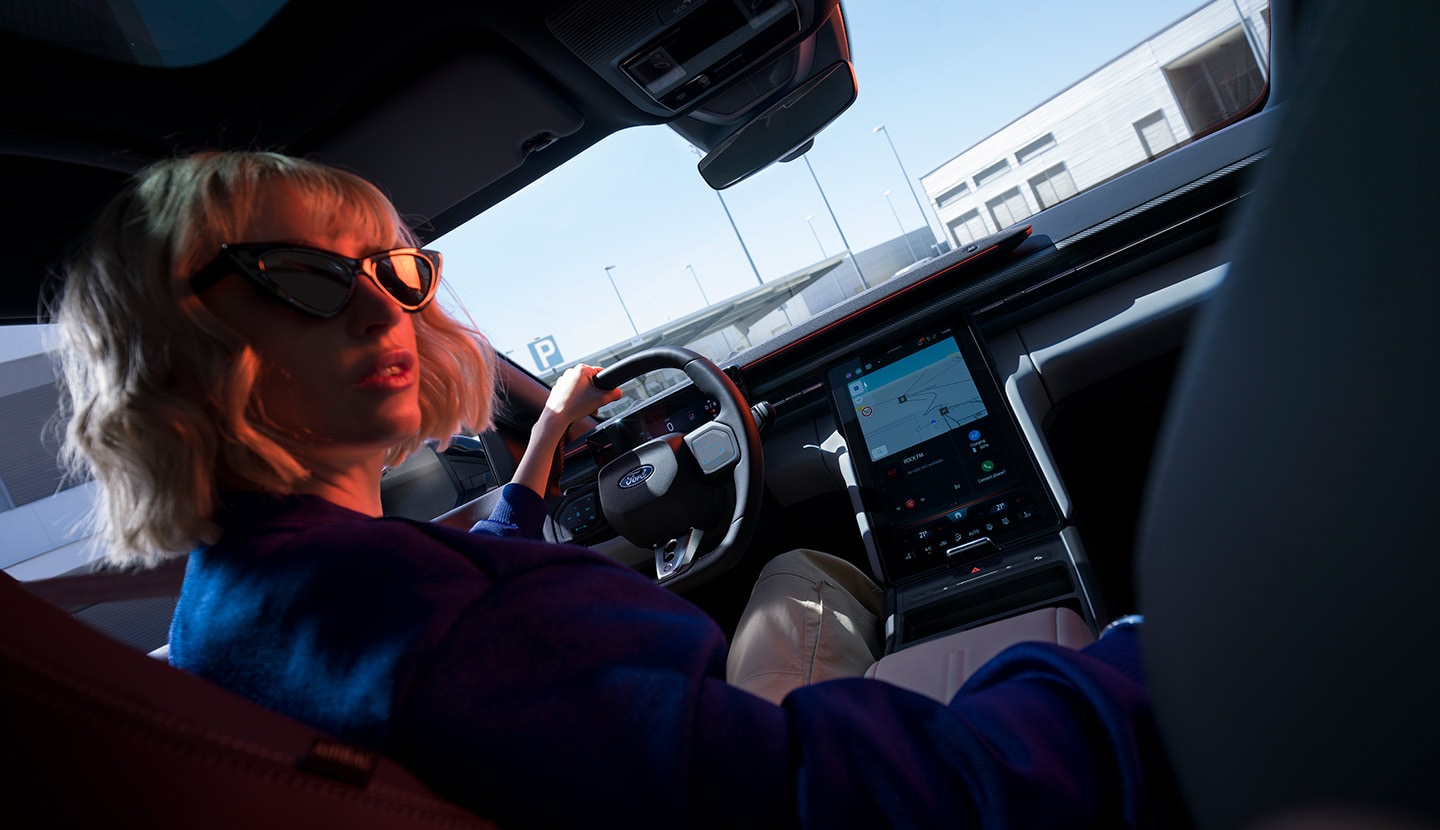 Een vrouw zit op de bestuurderszetel van een geparkeerde Ford Capri®, met zicht op het stuur en een groot SYNC® Move-touchscreen.