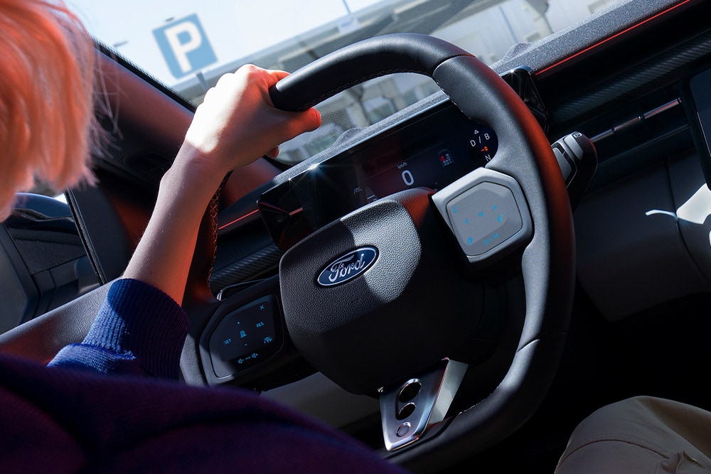 Een close-up van een vrouw die het stuur vasthoudt in een geparkeerde Ford Capri, met zich op het digitale instrumentenpaneel.