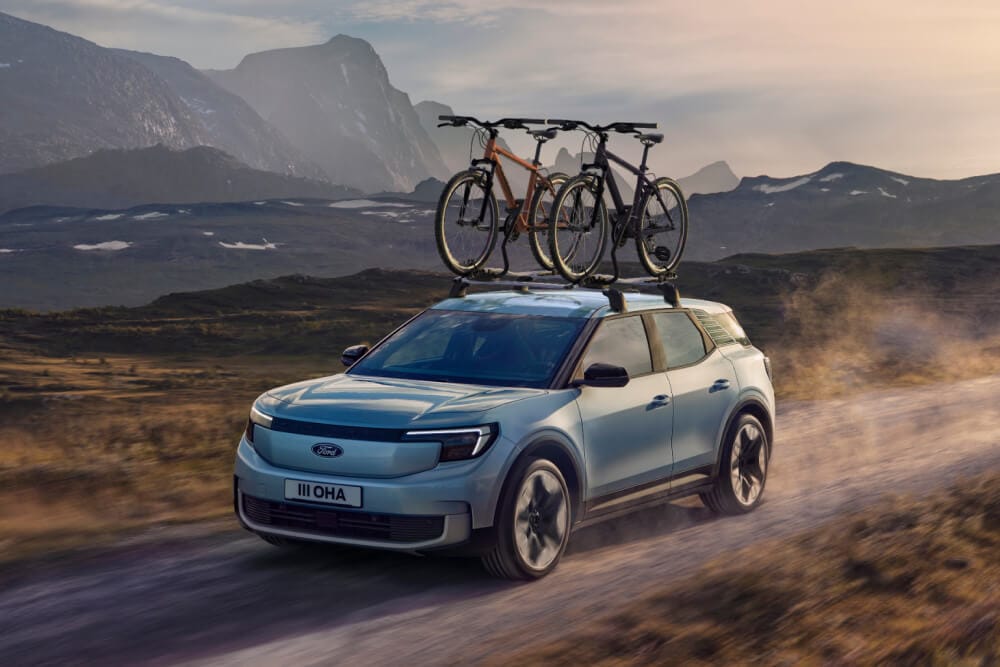 Blauwe Ford Explorer onderweg op een zandweg, met dakdragers, een fietsdrager en twee fietsen op het dak aangebracht.
