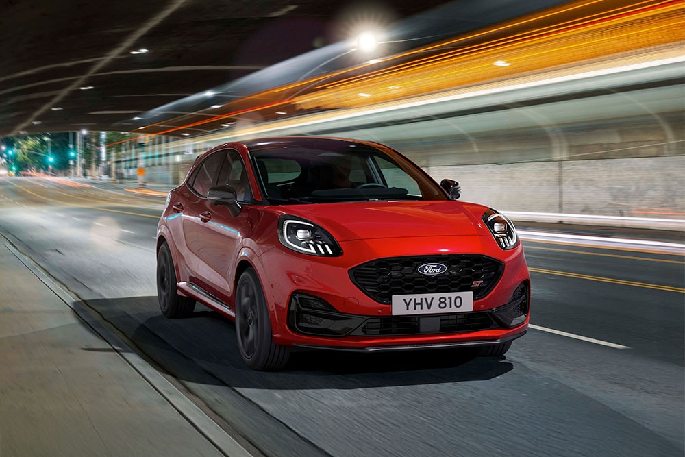 Een rode Ford Puma ST sportieve SUV rijdt in het donker over een fel verlichte stadsweg.