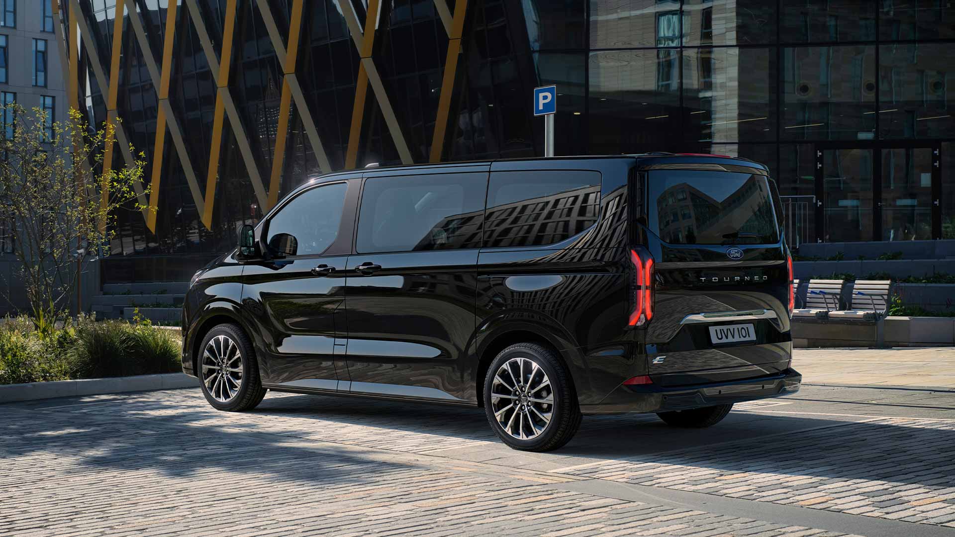 Een zwarte Ford E-Tourneo Custom staat geparkeerd met de zijkant en achterkant voor een modern gebouw.