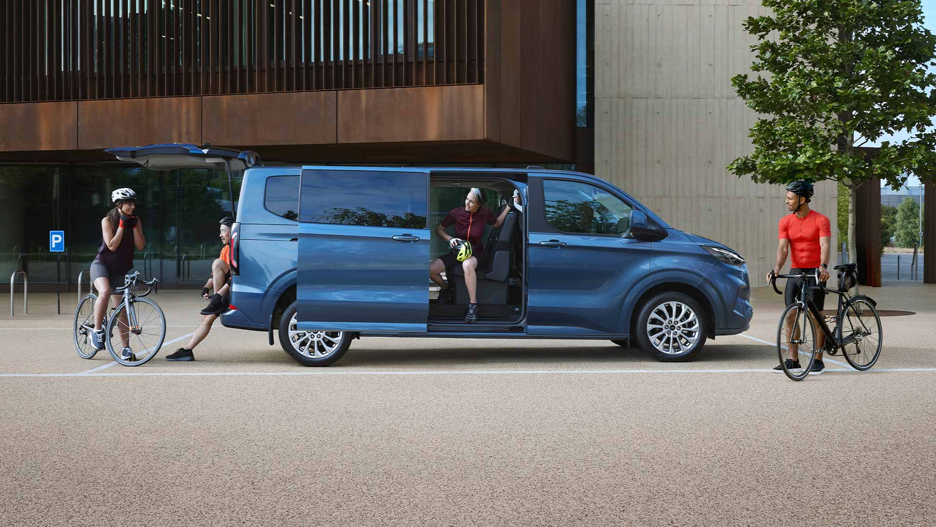 Blauwe Ford Tourneo Custom geparkeerd met zijn zijdeuren open en mensen met fietsen in de buurt. 
