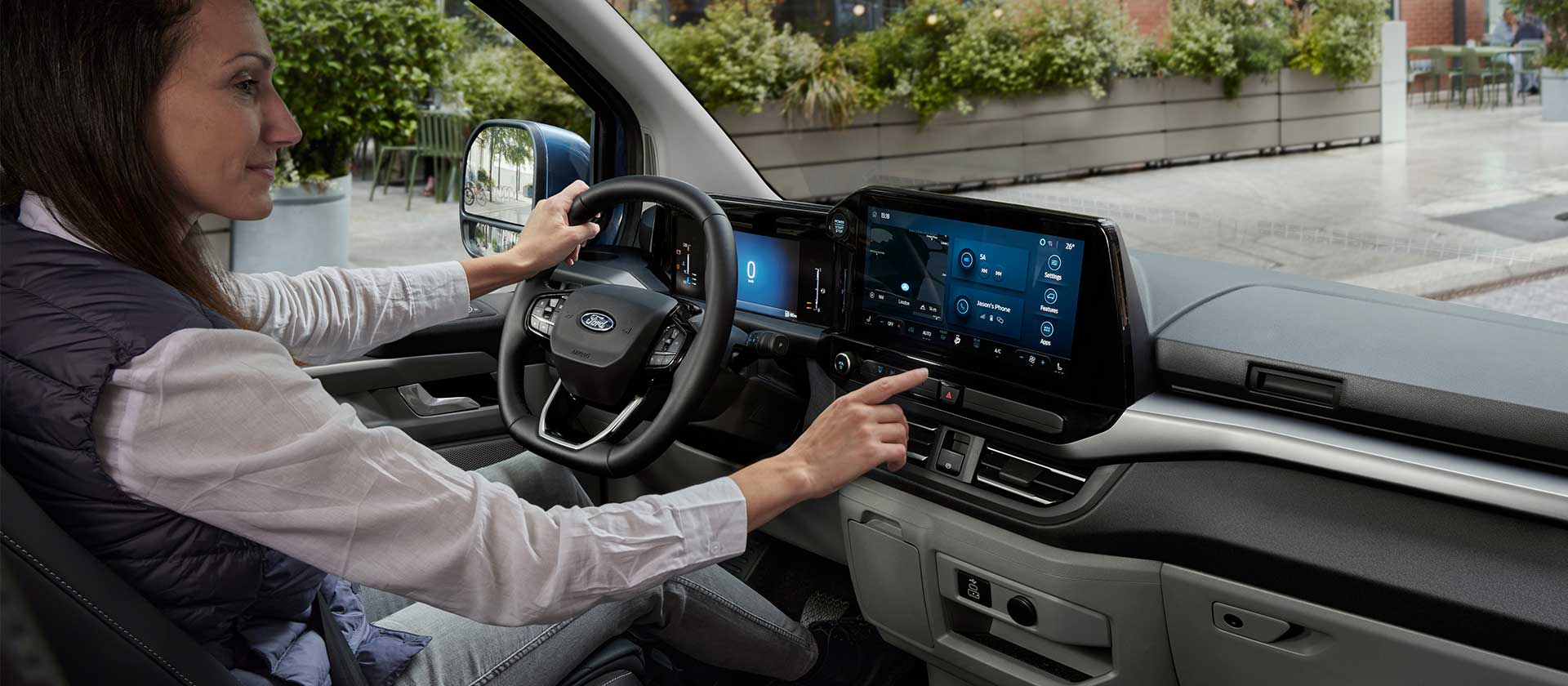 Een vrouw zit op de bestuurderszetel van de Ford E-Tourneo Custom en communiceert met het centrale touchscreen.