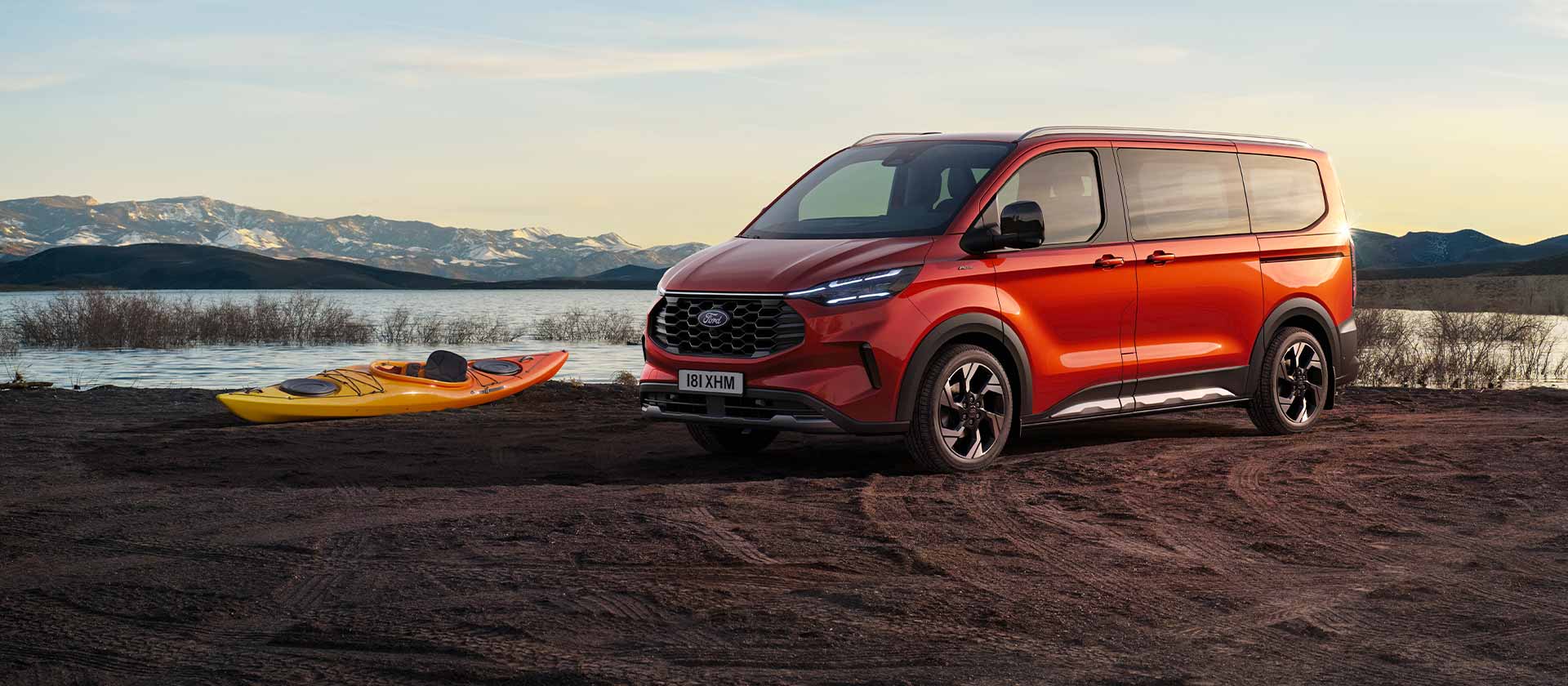 Een rode Ford Tourneo Custom bestelwagen staat geparkeerd bij een meer met een kajak.