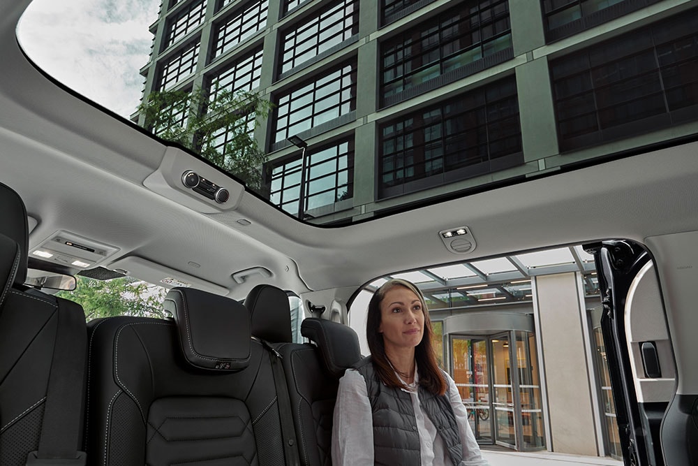 Een vrouw zit achterin de Ford Tourneo Custom en kijkt omhoog door het panoramische zonnedak.