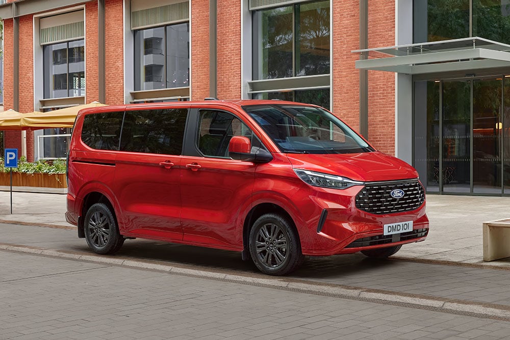 Een rode Ford Tourneo Custom staat geparkeerd in een stadsstraat voor een bakstenen gebouw
