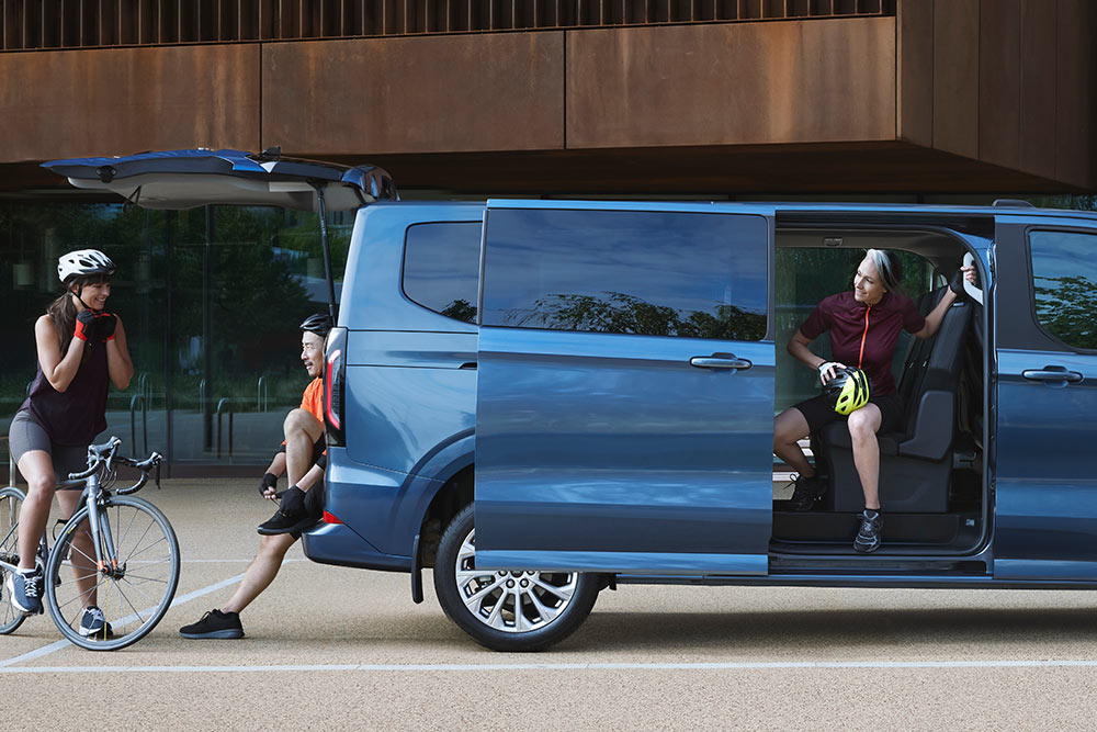 Blauwe Ford Tourneo Custom geparkeerd met zijn zijdeuren open en mensen met fietsen in de buurt. 
